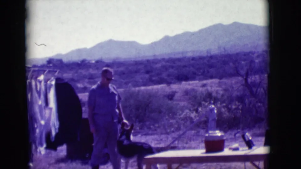 1966 Clarkdale, Arizona 8mm frame of a man in light clothing and sunglasses standing with a dark dog near laundry, a table, and mountains.
