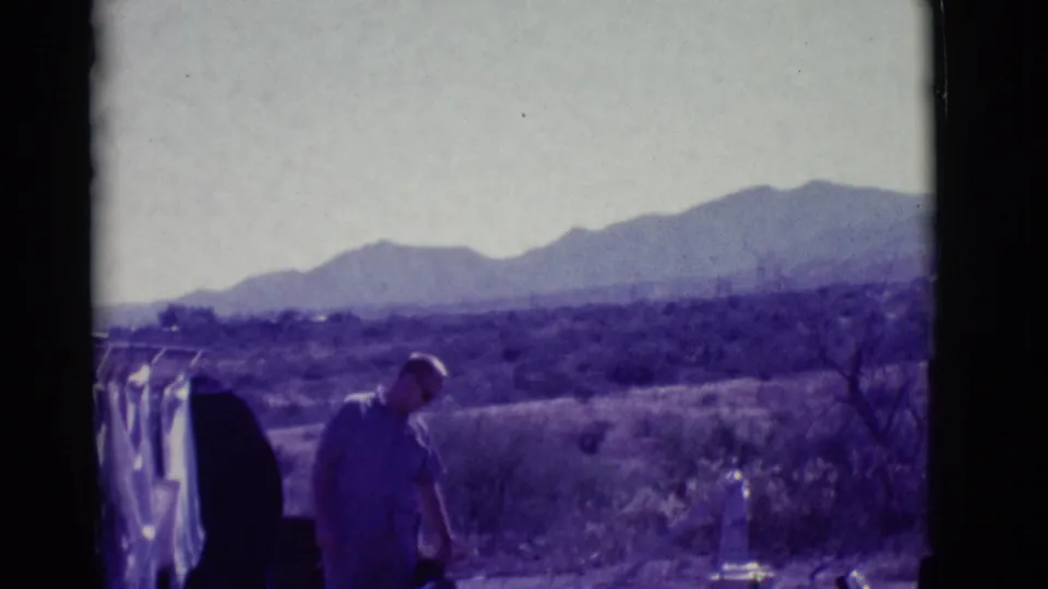 1966 Clarkdale, Arizona frame showing a man bending toward a dark dog near a clothesline, desert brush, and distant mountain ridges.
