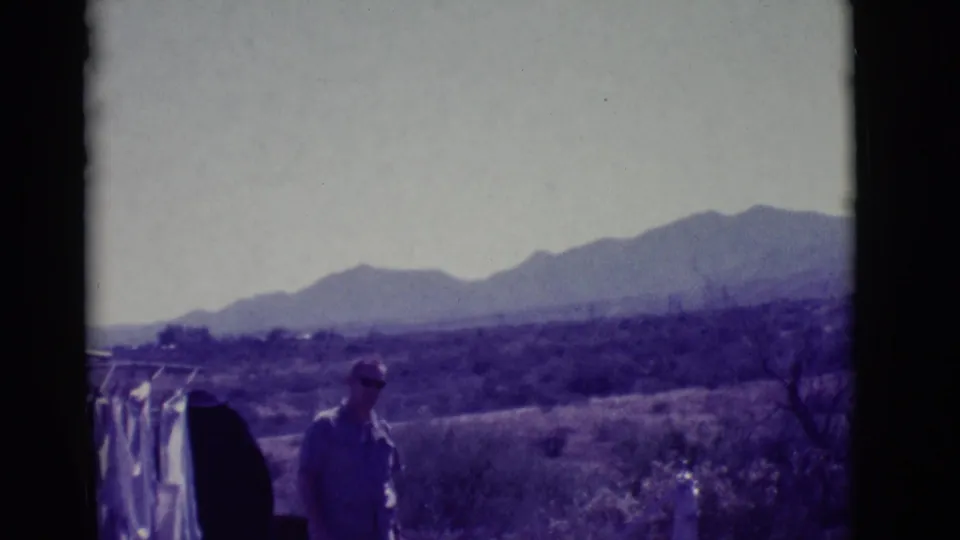 1966 Clarkdale, Arizona 8mm frame of a man standing near hanging laundry, scrubland, and hazy mountains under bright daylight.