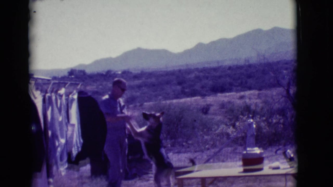 In the Clarkdale desert setting, the man faces a German Shepherd near hanging laundry, with a table and cooler at the right edge.