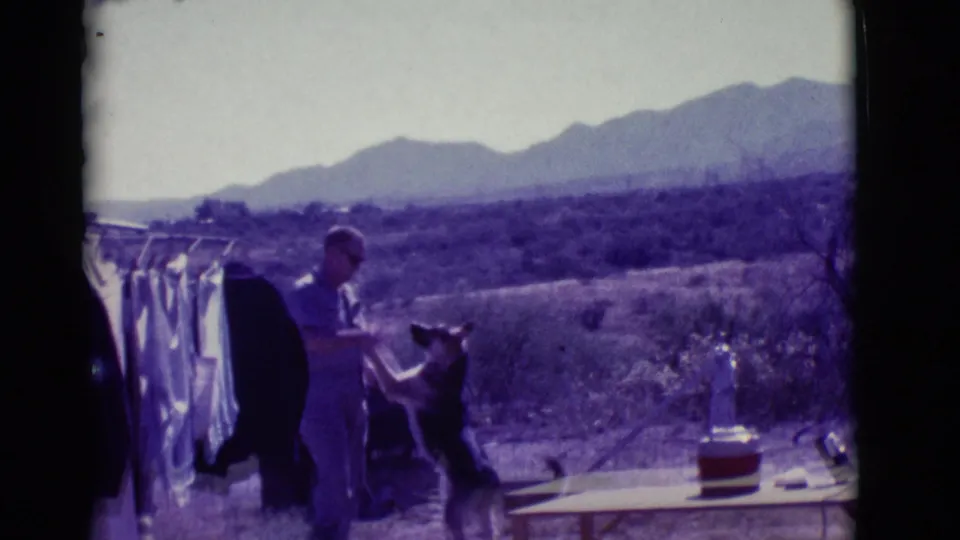 In the Clarkdale desert setting, the man faces a German Shepherd near hanging laundry, with a table and cooler at the right edge.