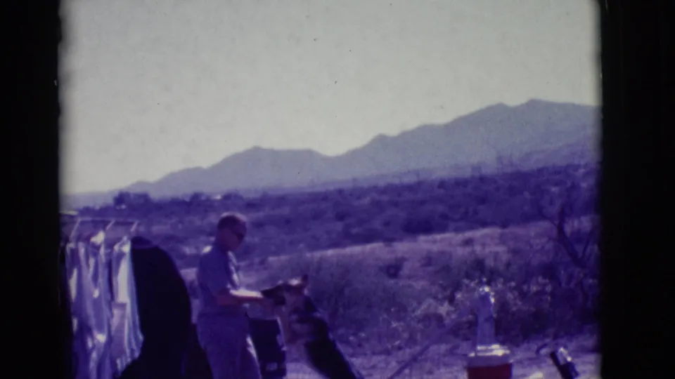 The man leans toward the German Shepherd as it reaches up, with laundry, a cooler, scrubland, and distant mountains visible around them.
