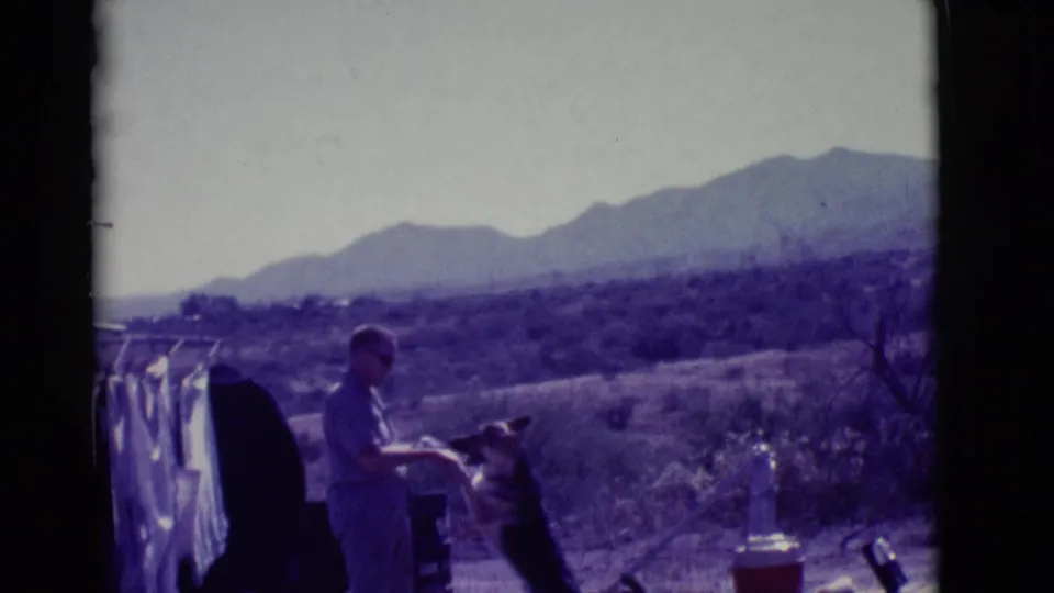 In 1966 Clarkdale, Arizona home-movie footage, a man in sunglasses interacts with a German Shepherd near a clothesline with mountains beyond.