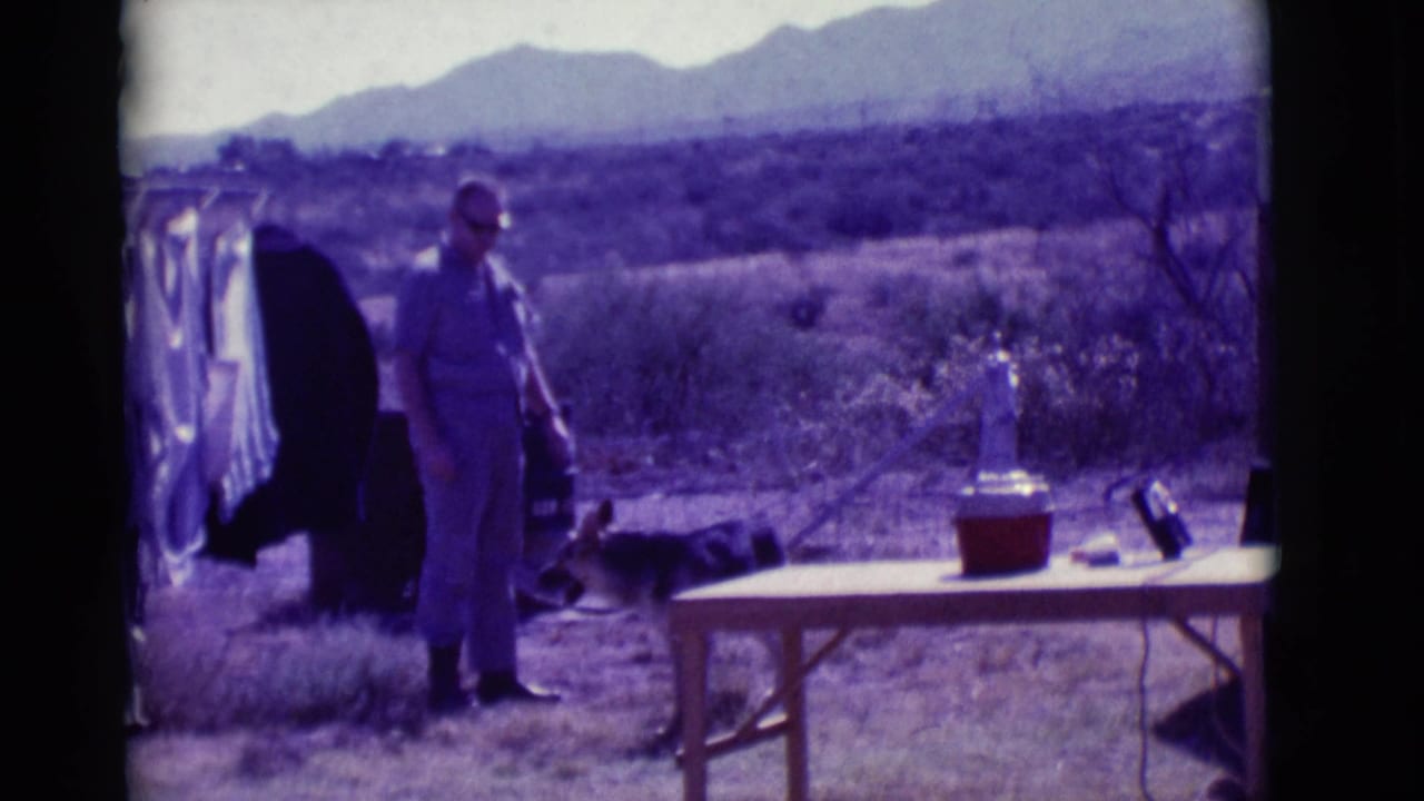 Man stands near a clothesline, folding table, red cooler, and dog in a desert landscape.