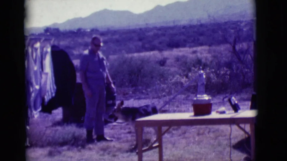 A man stands in a Clarkdale desert yard near a German Shepherd, folding table, red cooler, clothesline, and distant mountains.