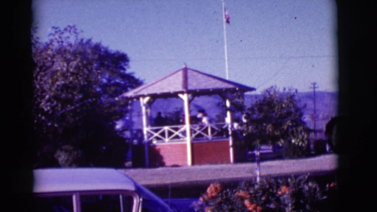 1966 Clarkdale, Arizona park view with a red brick gazebo, people gathered inside, flagpole, trees, distant hills, and car edge.