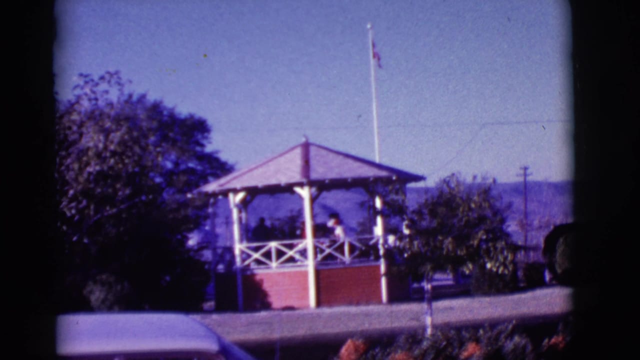 1966 Clarkdale, Arizona gazebo scene with people inside the structure, a flagpole rising behind it, trees, hills, and flowers in front.