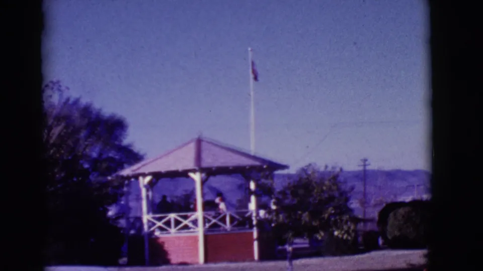 1966 Clarkdale, Arizona park gazebo with people under the roof, a flagpole above, blue sky, trees, hills, and a car roof edge.
