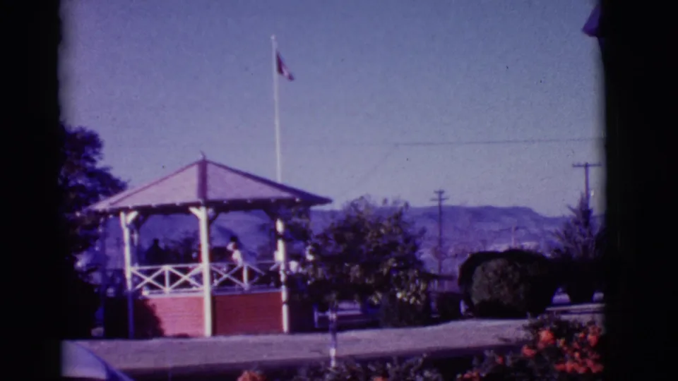 1966 Clarkdale, Arizona park shot showing people in a gazebo, flagpole behind, trees, distant hills, flowers, and film frame edge.