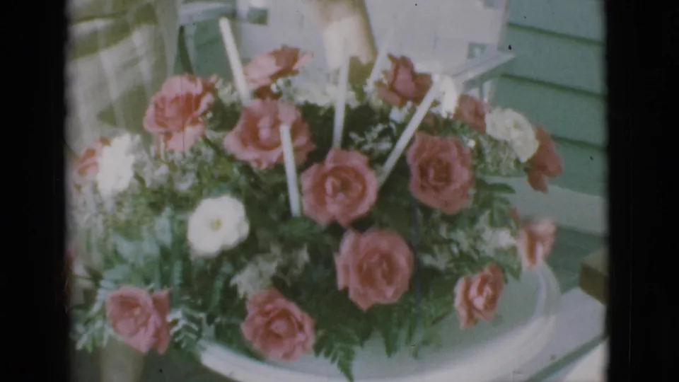 1966 Clarkdale, Arizona frame showing pink roses, white flowers, ferns, and candles arranged on a white table by green siding.