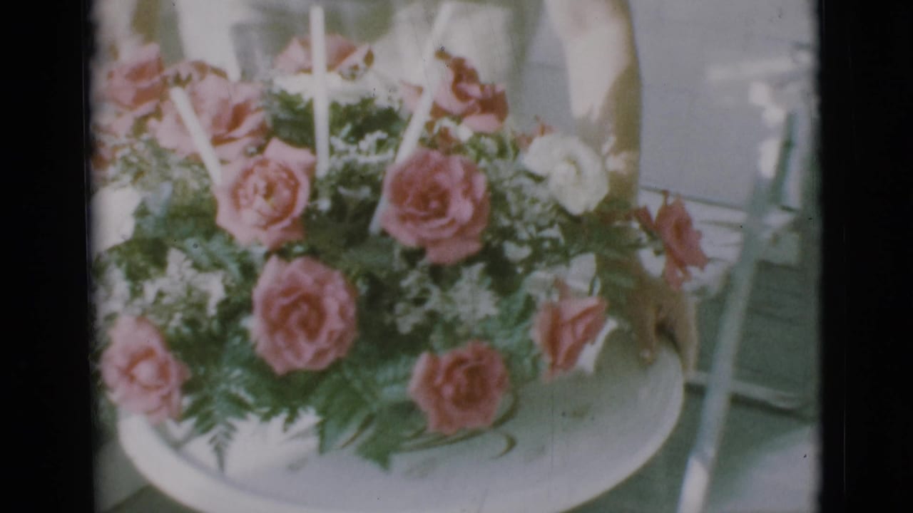 1966 Clarkdale home-movie frame of roses and greenery on a tray, with part of a light dress at the edge.