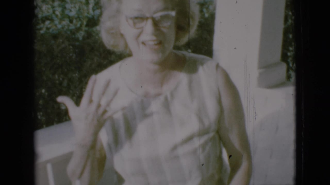 Woman in glasses smiles on a porch, gesturing with one hand beside a railing.