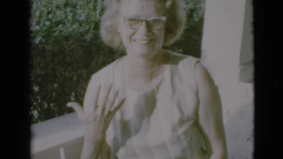 A 1966 home-movie frame shows the woman smiling, with one hand lifted near the porch railing and leafy background.
