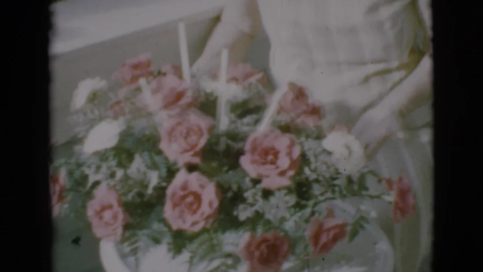 1966 Clarkdale home-movie frame showing hands arranging a pink and white rose bouquet on a porch.