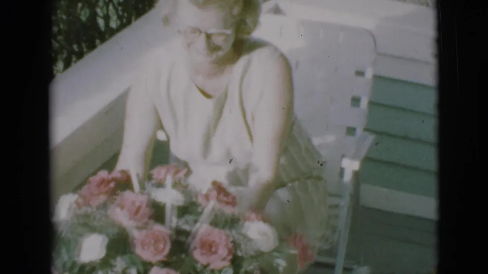 A woman seated on a porch leans toward a pink and white rose bouquet in 1966 Clarkdale home-movie footage.