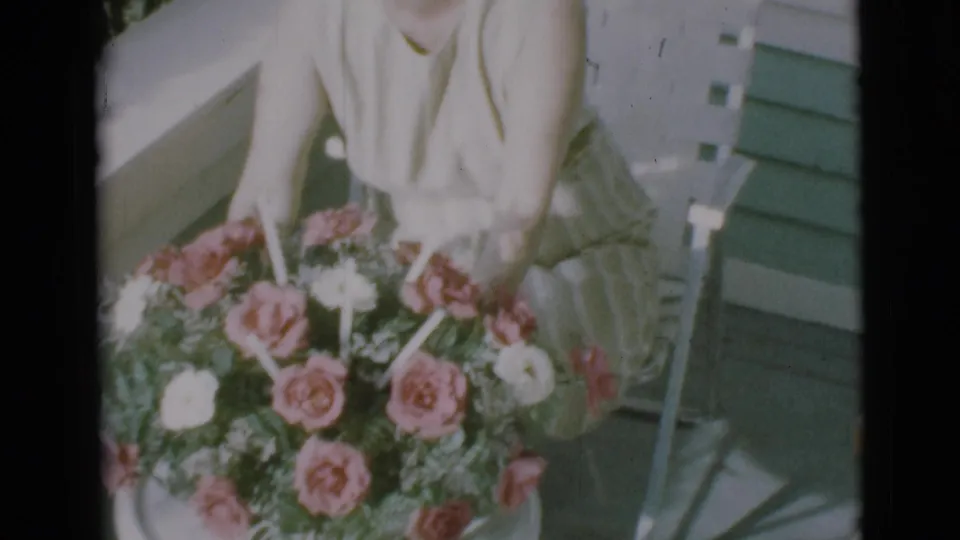 The woman reaches into a large rose bouquet beside a porch chair in the 1966 Clarkdale home-movie scene.
