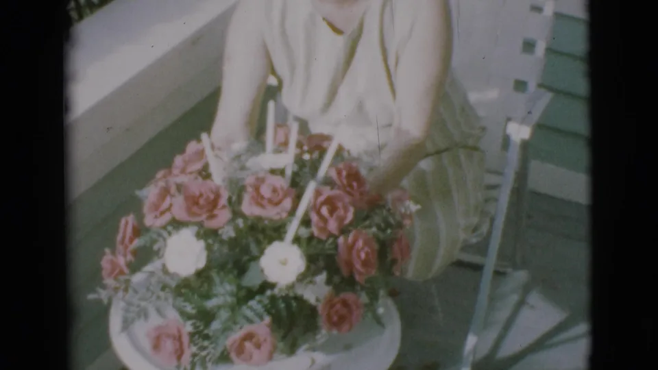 The woman turns the small round table, showing the rose arrangement and candles in soft 1966 8mm color.