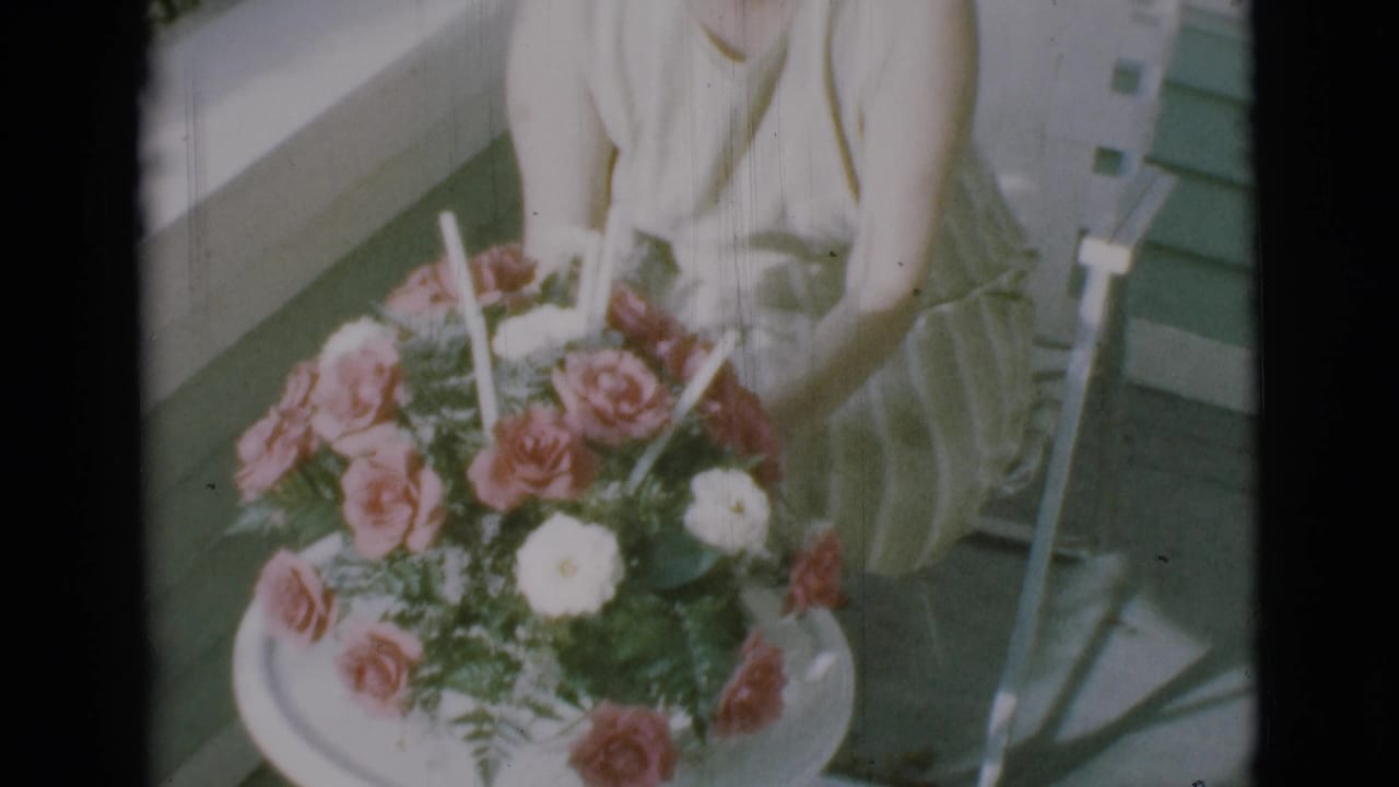 Woman seated in a lawn chair beside a large bouquet of pink and white flowers on a porch.