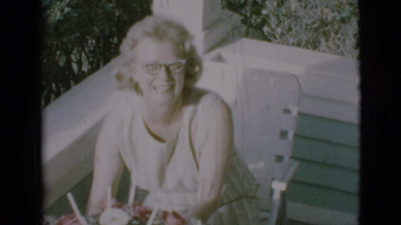 Woman in glasses sits on a porch smiling behind a large floral arrangement.