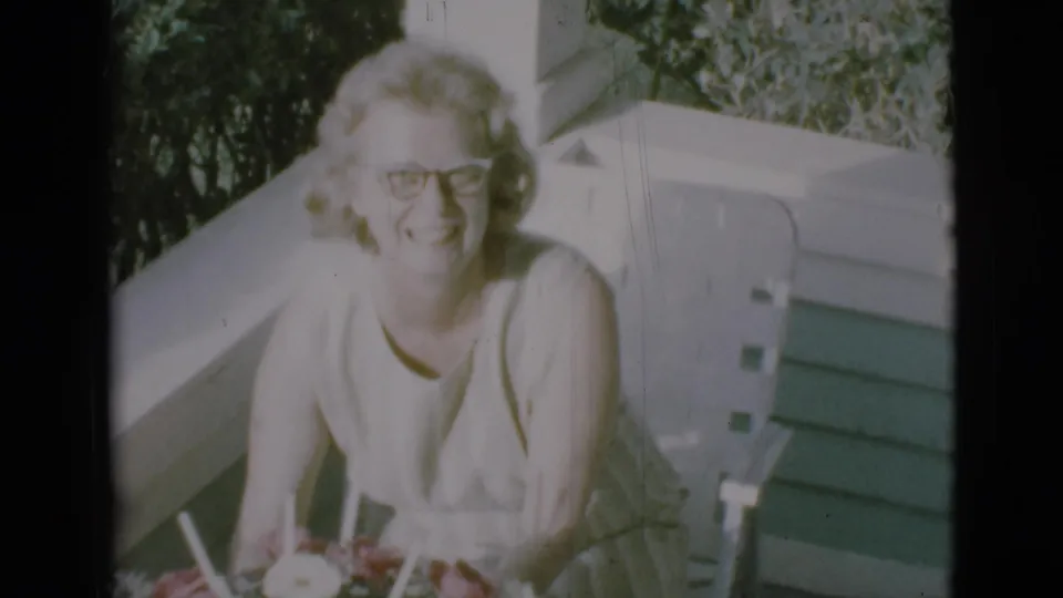 Adult woman in glasses smiles toward the camera on a Clarkdale porch in 1966, with flowers in front of her.