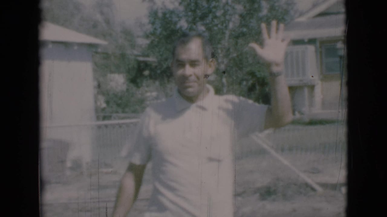 Man in a light polo shirt waves outdoors near fencing and trees.
