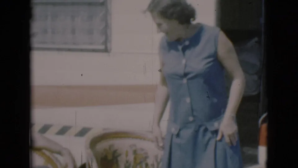 In Clarkdale, Arizona in 1966, a woman in a blue dress stands near a camper doorway and wicker-style lawn chair.