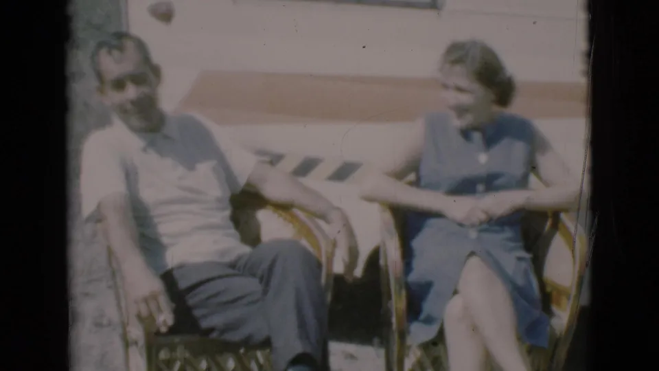 A seated man and woman relax beside a camper, with the woman turned toward him in the 1966 home-movie frame.