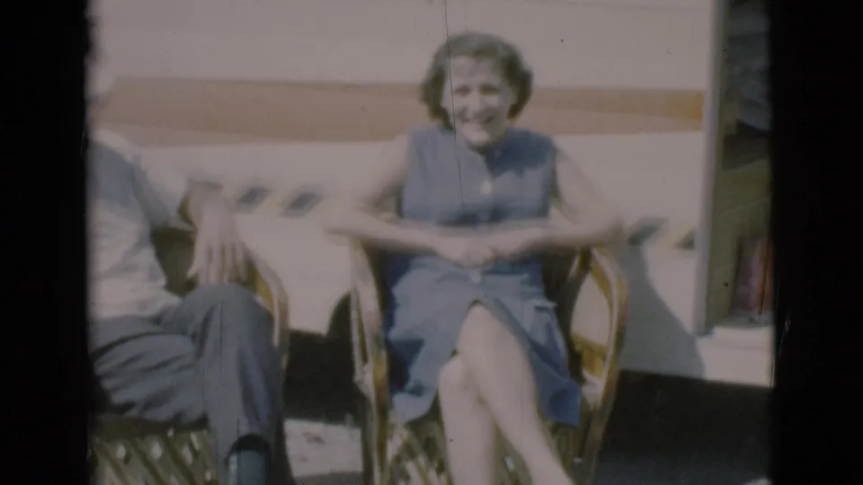 The woman sits facing the camera in a blue sleeveless dress, with the man partly cropped beside her outside the caravan.