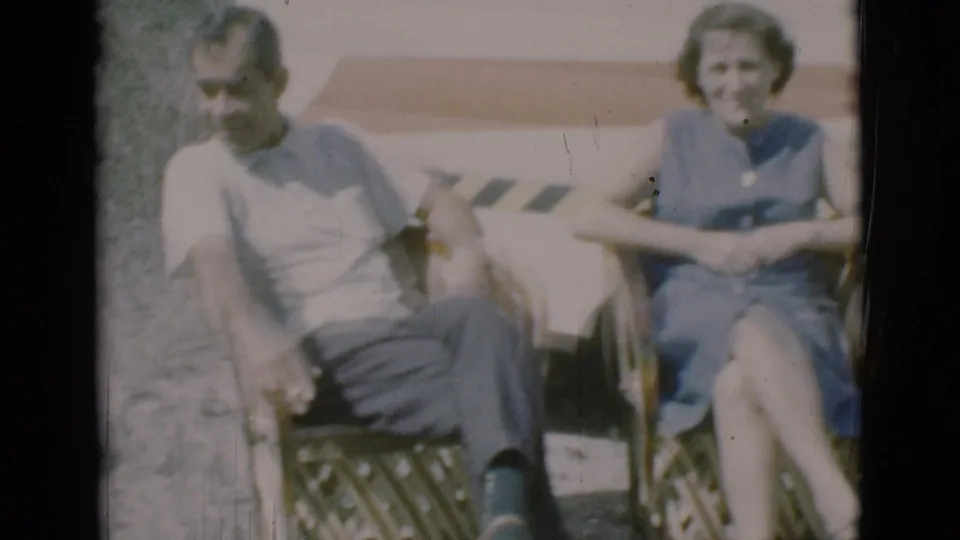 The adult couple sits together in wicker chairs outside the caravan, smiling in faded 1960s 8mm color.