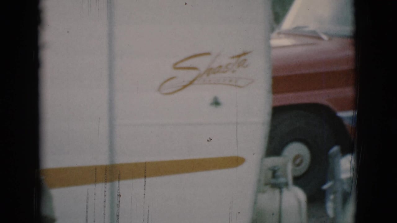 White Shasta trailer with gold stripe, propane tank, and red vehicle partly visible.