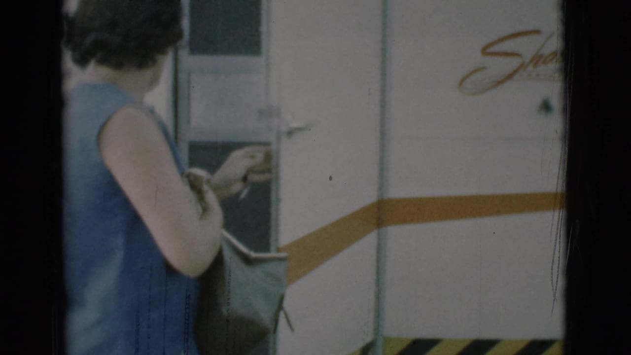 A woman in a sleeveless blue dress stands beside the Shasta trailer door in Clarkdale, Arizona, with a bag at her side.