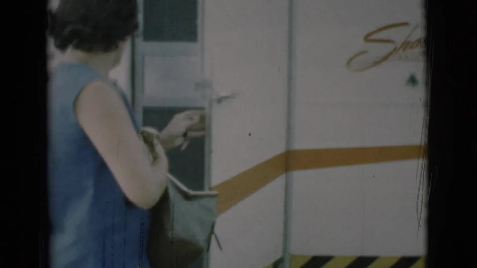 A woman in a sleeveless blue dress stands beside the Shasta trailer door in Clarkdale, Arizona, with a bag at her side.