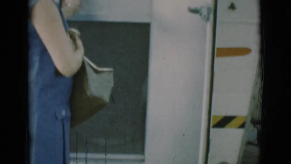 Close 8mm frame of the woman near the trailer doorway, with her bag and the white trailer wall filling most of the view.