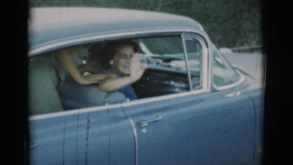 The woman smiles toward the camera and raises her hand in a wave from the blue vintage car in the home-movie frame.