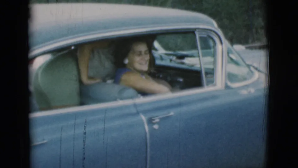 The woman remains seated in the blue car, smiling toward the camera as the brief farewell moment ends.