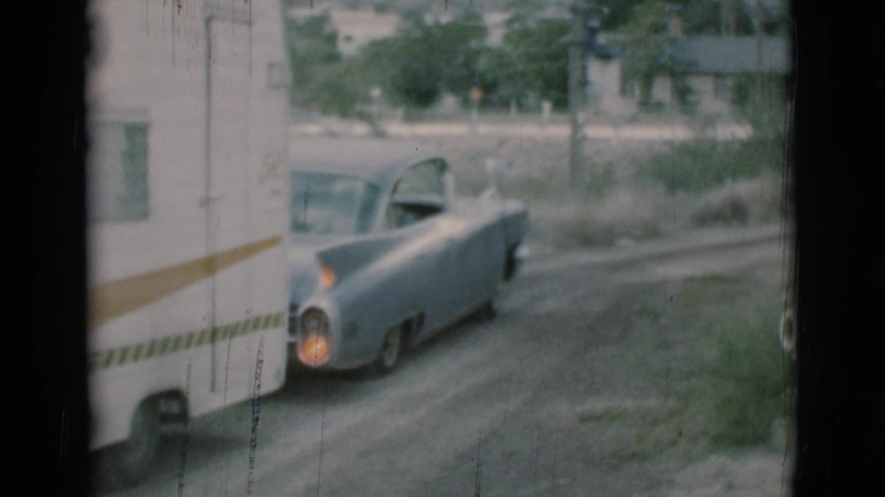 Light blue car towing a white travel trailer along a dirt road near trees and a building.