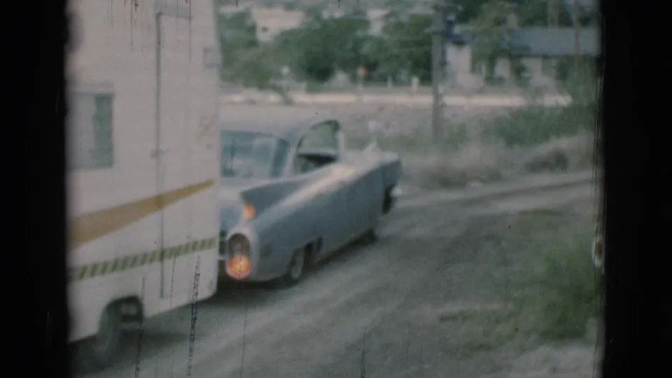 The light blue car pulls a white travel trailer along a rural dirt road in Clarkdale, Arizona, with trees and a small building in the background.