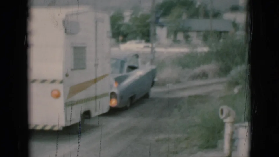 A white travel trailer follows the light blue car along the dirt road, with roadside grass, a small building, and soft 8mm texture visible in the frame.