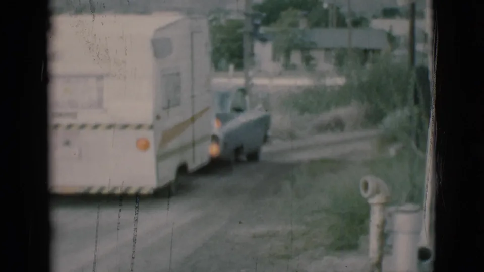The car and white travel trailer continue away along a rural Clarkdale road, framed by grass, trees, a small building, and a pipe or post in the foreground.