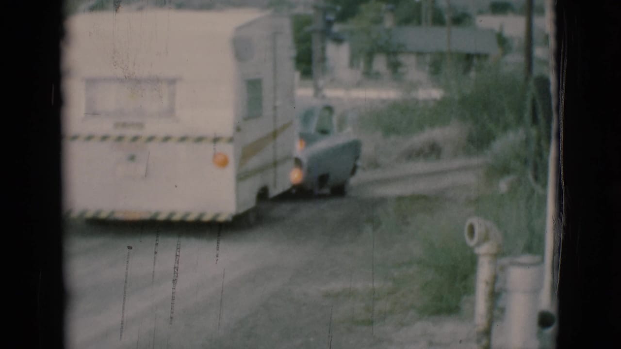 White caravan trailer towed by a gray sedan on a dirt road, seen through film gate edges.