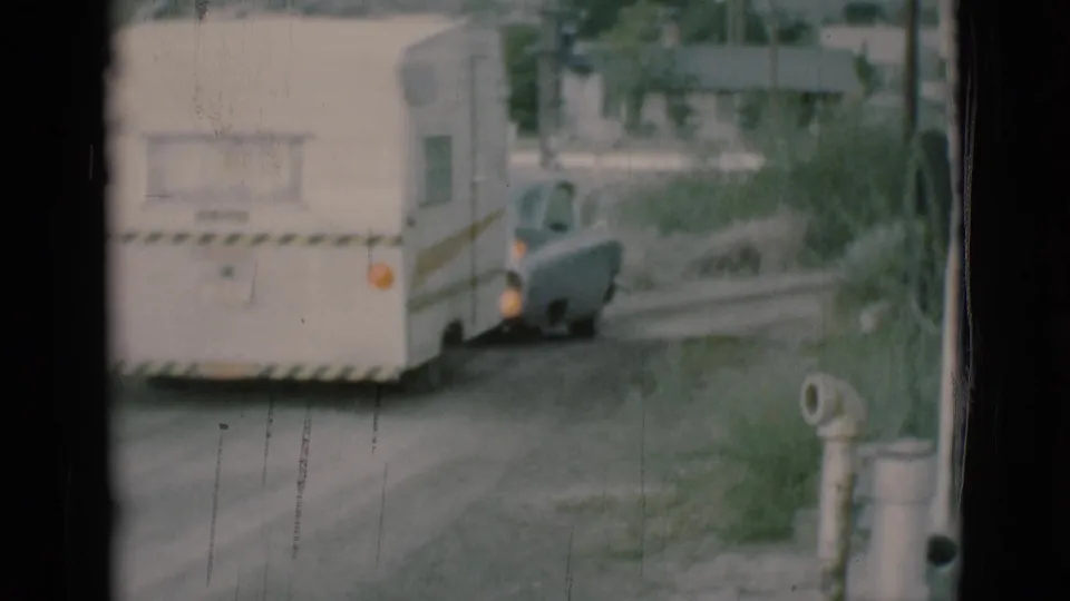 1966 Clarkdale, Arizona home-movie frame showing a white caravan trailer and gray sedan moving away on a dirt road.