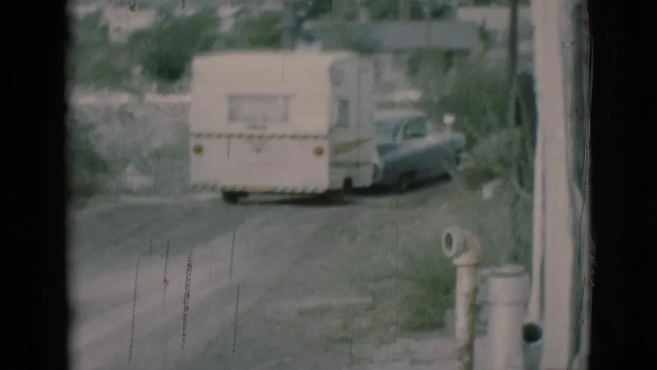 The trailer and towing sedan appear farther down the dirt road, framed by 8mm film edges and roadside details.
