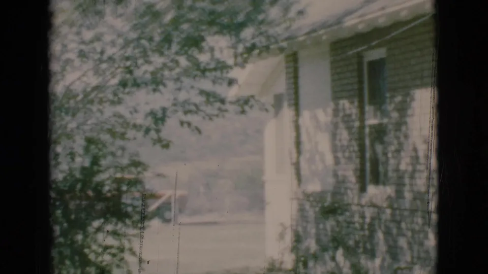 1966 home-movie frame of a Clarkdale house side with dark shingle siding, a porch railing, and branches in the foreground.