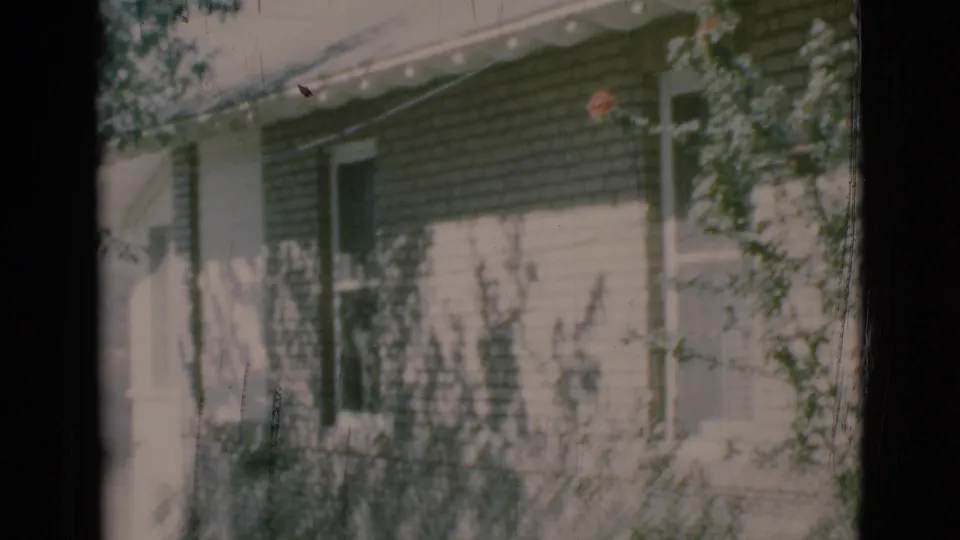 1966 8mm frame from Clarkdale, Arizona showing a brick house side exterior with white trim, roof eaves, shrubs, and soft tree shadows.