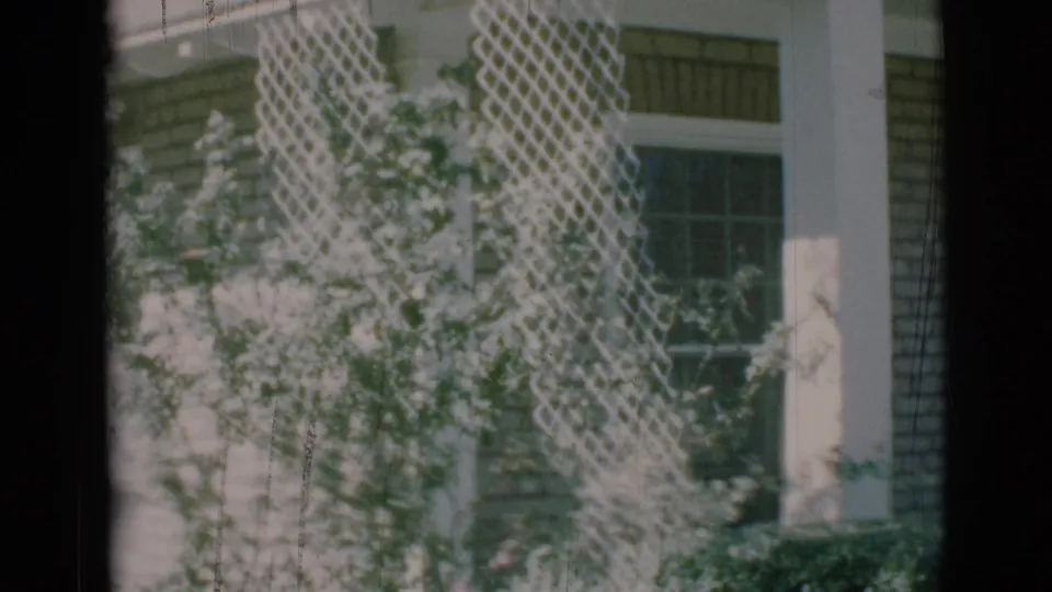 1966 8mm frame from Clarkdale, Arizona showing white latticework, a window, brick siding, and shrubs during the house exterior pan.