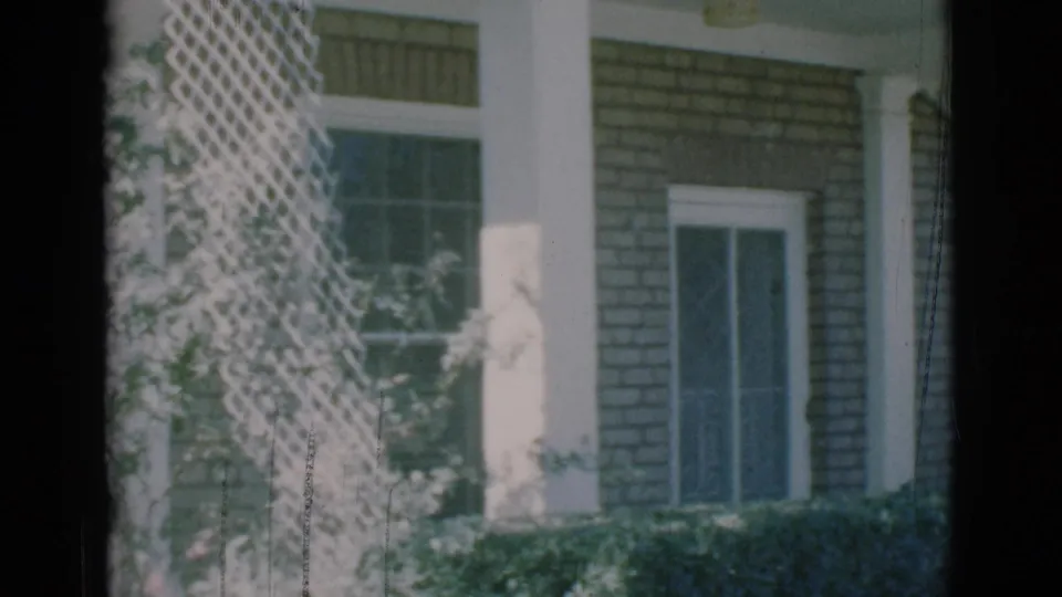 1966 8mm frame from Clarkdale, Arizona showing the front portion of a brick house with white trim, windows, latticework, and a low hedge.