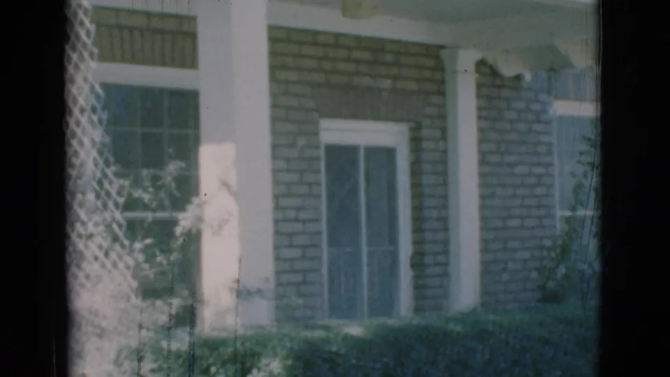 1966 Clarkdale, Arizona home-movie frame showing a brick house facade with white columns, window screens, and hedges seen from behind a screen.
