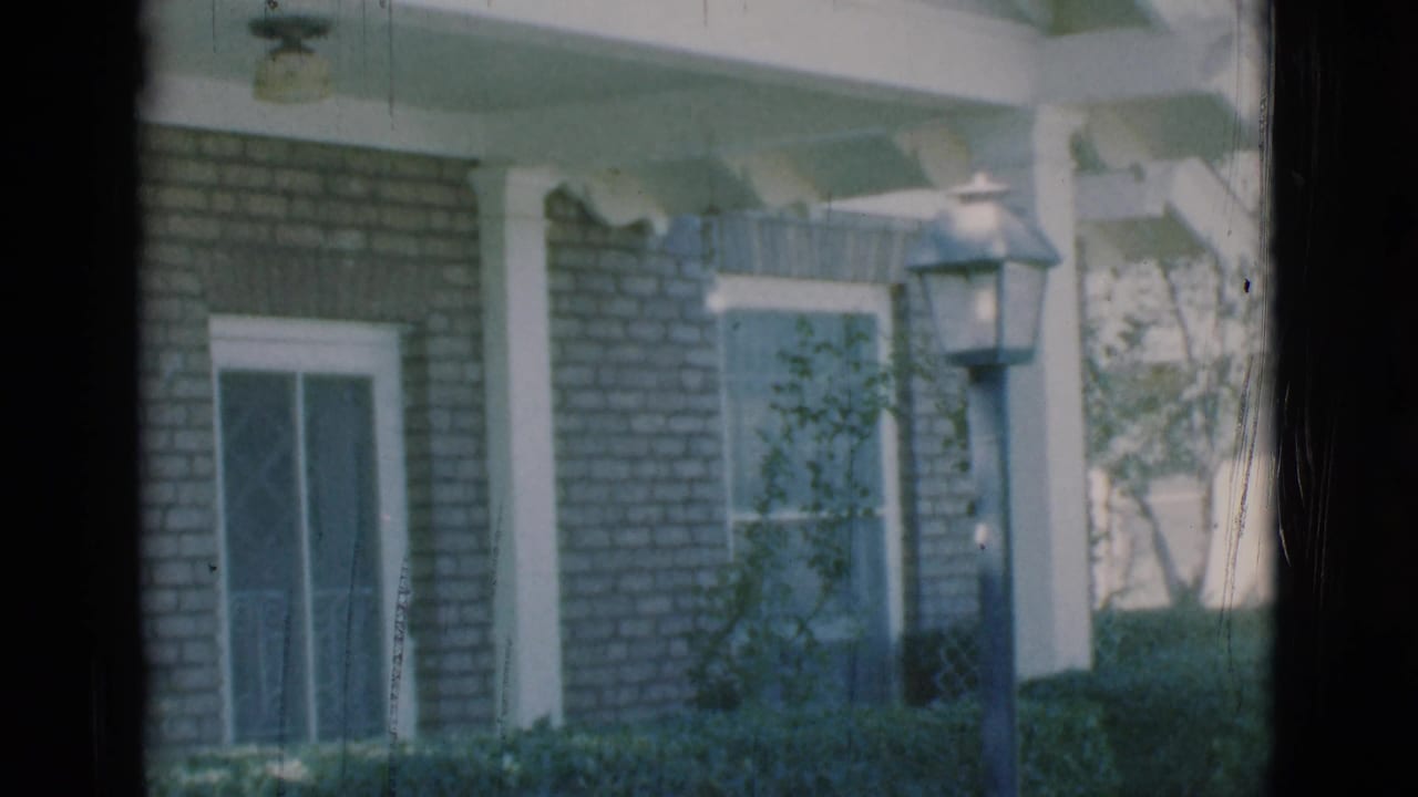 Brick house front with white columns, window awnings, hedges, and a yard lamppost near the porch.
