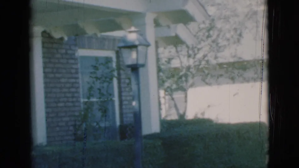 1966 Clarkdale, Arizona home-movie view of a brick house corner with a screened window, white porch support, hedges, and a yard lamp.