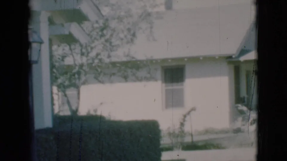 1966 Clarkdale, Arizona frame showing the side of a light-colored house with a small window, hedge border, lawn, and walkway.